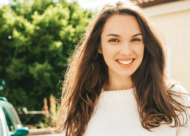 Smiling woman with long dark hair in white top, outdoor setting with greenery background.