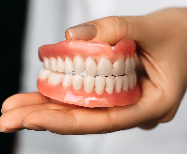Hand holding full set of dentures displaying artificial teeth against white background.