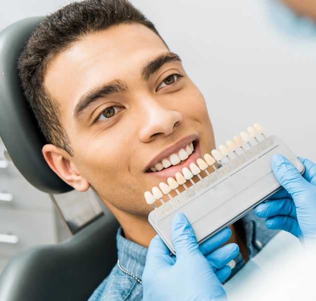 Man in dental chair smiling while dentist holds tooth shade guide for color matching.