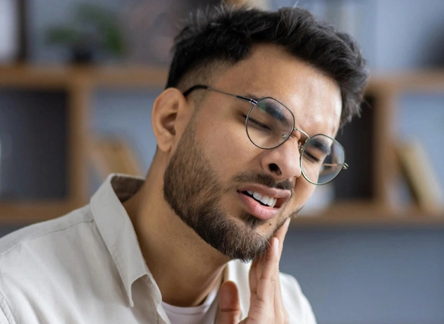 Man wearing glasses touching cheek in pain, modern dental office interior background