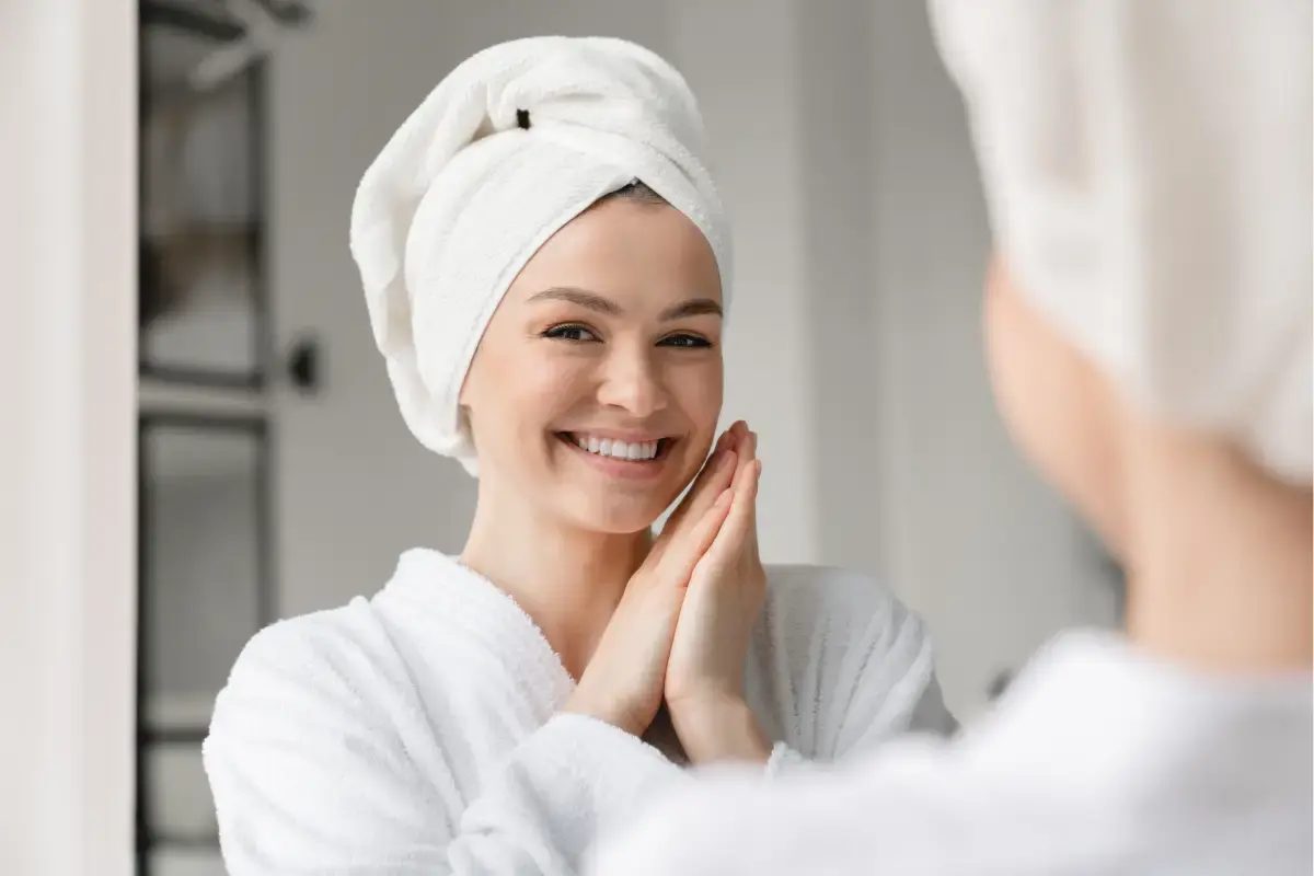 Smiling woman admiring her bright, healthy teeth in bathroom mirror after whitening treatment.